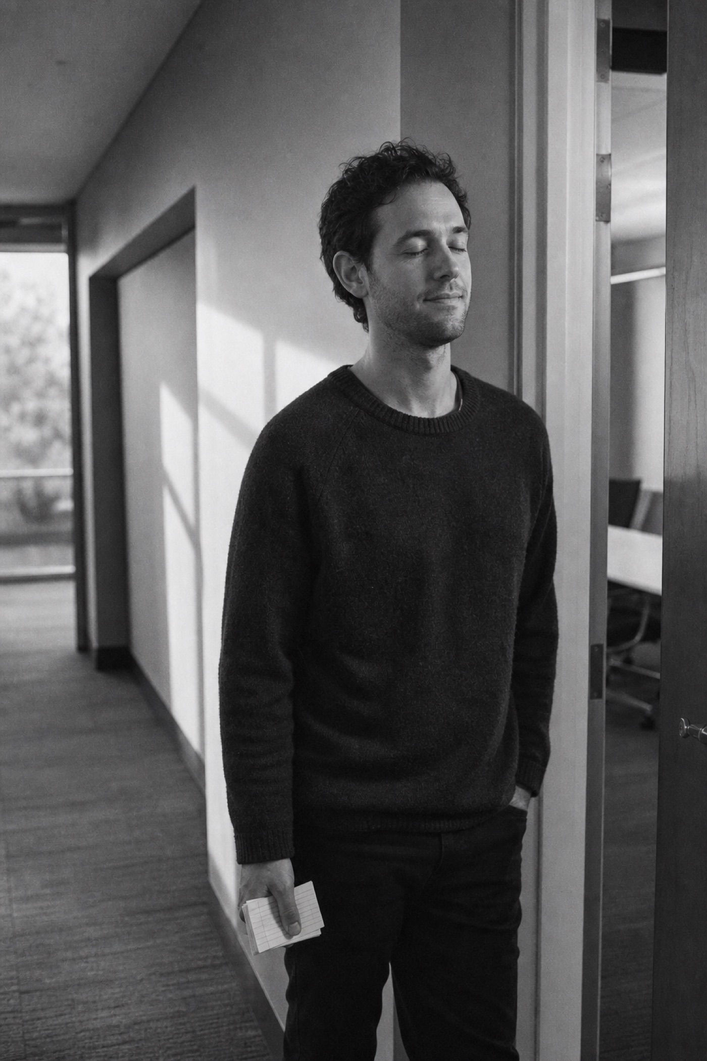 Black and white photo of a man pausing quietly in a hallway, eyes closed, notes in hand, taking a breath before rehearsing.