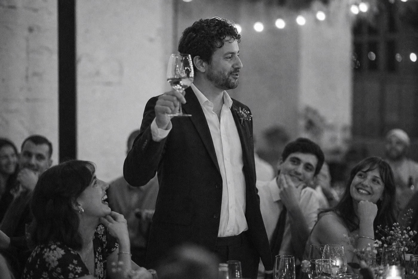 Black and white photo of a man raising a toast at a wedding reception while guests around him smile.